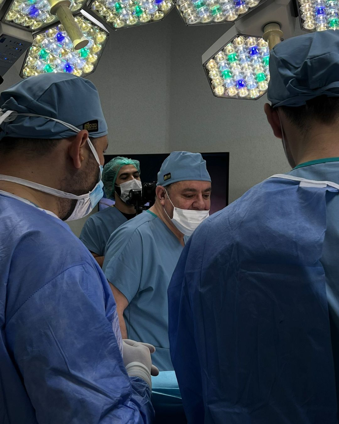 Surgeons in blue scrubs and masks performing surgery under bright overhead lights in an operating room.