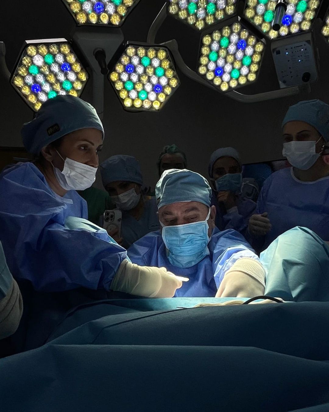 Surgeons in blue scrubs and masks performing surgery under bright overhead lights in an operating room.